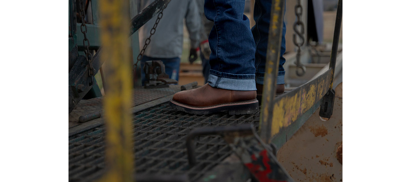 Un hombre que viste pantalones vaqueros con dobladillo y botas Homeland de color marrón está parado sobre una plataforma de rejilla de metal, con cadenas industriales y equipos visibles en el fondo.
